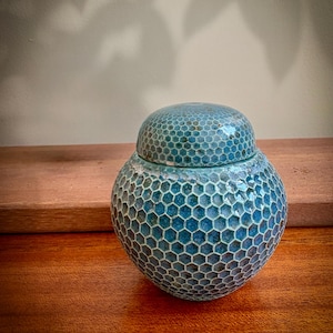 May include: A round ceramic jar with a lid, featuring a textured blue honeycomb pattern across its entire surface. The glaze displays variations of deep and light blue tones within each hexagonal cell, creating a detailed, tactile appearance.