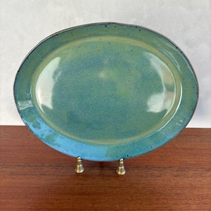 May include: An oval-shaped ceramic platter featuring a glossy glaze in shades of teal, turquoise, and light green. The surface has a subtle speckled texture throughout, with a darker rim defining the edge of the dish, supported by a small metal stand.
