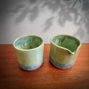May include: Two ceramic vessels with a smooth, glossy glaze in shades of pale green, soft yellow, and light blue. One vessel is a simple cylindrical cup, while the other features a small, integrated pouring spout on its rim.