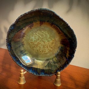 May include: A round ceramic bowl with a textured, uneven rim, featuring a glossy glaze that transitions from a light, mottled olive-green centre to deep, dark blue and brown tones along the edges, supported by a small, gold-coloured metal stand.