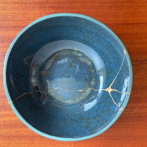 May include: A round ceramic bowl with a deep blue mottled glaze and a smooth interior surface. The bowl features a prominent crack repaired with gold-colored lacquer, creating a distinct branching pattern that extends across the base.