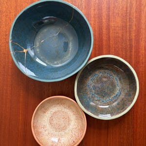Op de afbeelding: Drie ronde keramische kommen van verschillende groottes. De grootste kom is donkerblauw met een goudkleurige kintsugi-reparatielijn. De middelste kom heeft een gevlekt blauw en bruin glazuur. De kleinste kom heeft een lichtbruine en oranje afwerking.