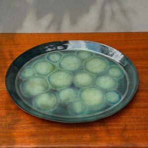 Può includere: Piatto ovale in ceramica con un motivo a nido d'ape verde e verde acqua. Il piatto ha un bordo verde acqua scuro e una finitura lucida. La superficie è un mix di verde chiaro e scuro, creando un aspetto strutturato. Il piatto è su una superficie di legno.