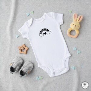 May include: White baby onesie with a black rainbow and heart design. The image includes a pair of striped baby shoes, a wooden star-shaped teether, and a plush bunny rattle. The items are arranged on a gray surface.