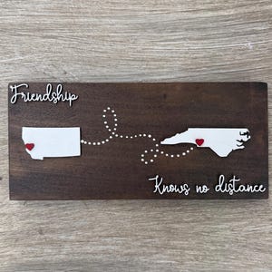 May include: Wooden sign with the words "Friendship Knows no distance". The sign features white cutouts of two state outlines, connected by a dotted line, each with a red heart. The background is a dark brown.
