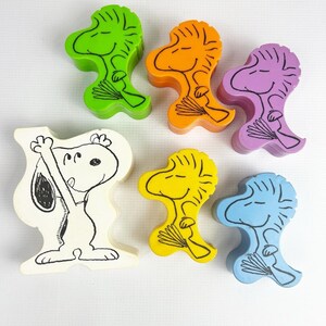 May include: A collection of colorful wooden figures featuring Snoopy and Woodstock characters. The figures are in various colors, including green, orange, purple, yellow, and blue. One figure is a black and white illustration of Snoopy.