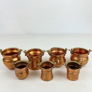 May include: A collection of seven small copper pots with brass handles arranged in two rows. The pots vary in shape, with some featuring rounded bodies and others having straight sides, all displaying a polished metallic finish.