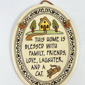May include: An oval ceramic wall plaque with a cream background and a black decorative border. It features a yellow house illustration, green trees, and a brown cat. The text reads: THIS HOME IS BLESSED WITH FAMILY, FRIENDS, LOVE, LAUGHTER, AND A CAT.