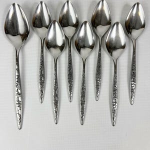 May include: A collection of eight silver spoons. Each spoon has a teardrop-shaped bowl and a textured handle. The spoons are arranged on a white surface, with the handles pointing downwards.