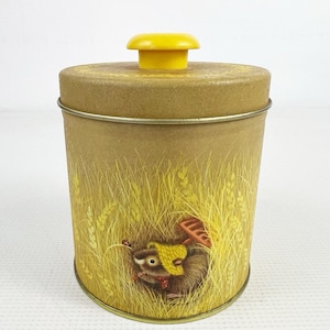 May include: A cylindrical tan metal canister with a bright yellow knob on the lid. The exterior features a printed illustration of a small brown animal wearing a yellow hat, surrounded by tall yellow wheat stalks and a small red ladybug.