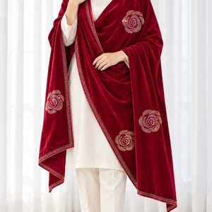 May include: A deep red velvet shawl featuring a border of small gold-toned studs and several large, circular rose-shaped embellishments made of shimmering crystals. The shawl is draped over a plain white long-sleeved tunic and white trousers.