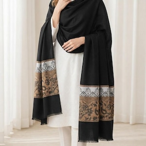 May include: A black shawl featuring a wide border with a tan and brown paisley pattern and a grey geometric band. The shawl has a fringed hem and is draped over a white long-sleeved tunic and white trousers, creating a layered appearance.