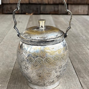 May include: A silver sugar bowl with an ornate design and a handle. The bowl has a lid and is made of metal.