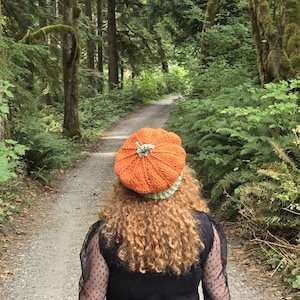 May include: A crocheted pumpkin-shaped hat in bright orange with a light green stem and ribbed texture, worn on the head. The hat features a rounded, segmented design that mimics the appearance of a pumpkin, with a fitted light green brim.