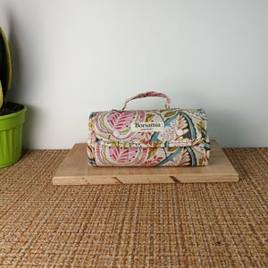 Puede incluir: Una bolsa de viaje rectangular con estampado floral, con tonos rosas, verdes y crema. Tiene un asa y el texto "Borsamia Handmade". La bolsa está colocada sobre una tabla de madera. La bolsa parece ser de tela.