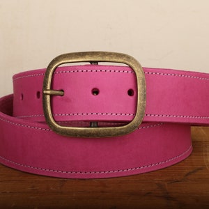 May include: A pink leather belt with a brass buckle. The belt is folded in half and the buckle is in the center of the image.