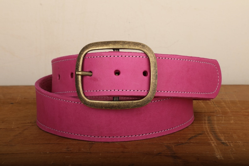 May include: A pink leather belt with a brass buckle. The belt is folded in half and the buckle is in the center of the image.
