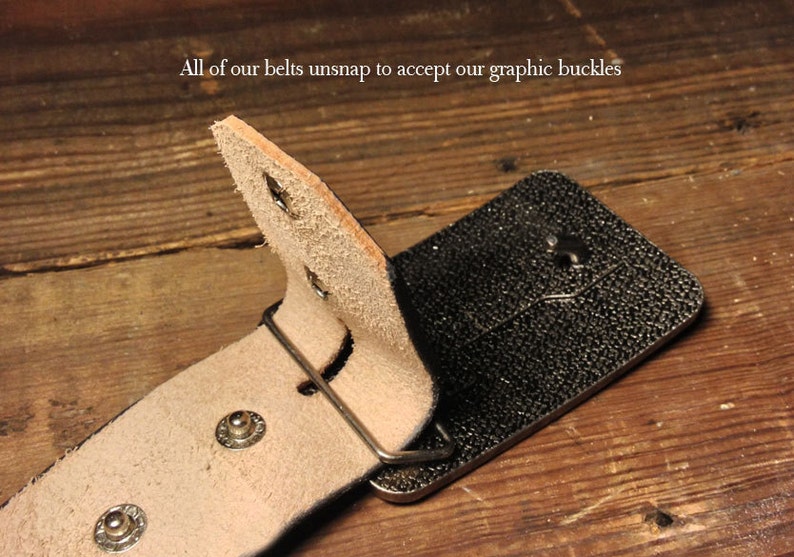 May include: A close-up of a brown leather belt with a black buckle. The belt is designed to unsnap to accept a graphic buckle. The text "All of our belts unsnap to accept our graphic buckles" is visible in the image.
