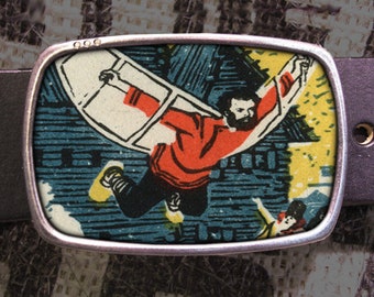 May include: A silver belt buckle featuring a colourful image of a person flying through the air. The person is wearing a red shirt and white trousers. The background is a blue and yellow pattern.