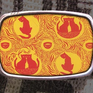 May include: A silver belt buckle with a yellow and red design featuring cats and bowls. The design is a repeating pattern of cats and bowls on a yellow background with red accents.