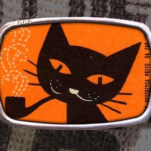 May include: A silver belt buckle with an orange background and a black cat smoking a pipe. The text "DYSTRYBICA PBZEC. 64 719" is on the right side of the buckle.