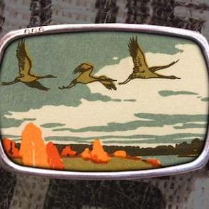 May include: A rectangular belt buckle with a silver-colored frame. The buckle features a landscape scene with three birds in flight over a body of water and orange-colored trees. The belt is black.