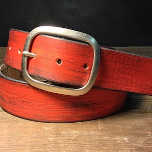 May include: A red leather belt with a silver-colored square buckle. The belt has a distressed finish with black streaks, and is rolled up on a wooden surface. The belt is approximately 1.5 inches wide.