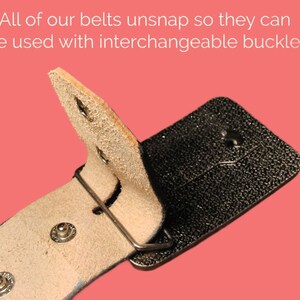 May include: A close-up of a brown leather belt with a black metal buckle. The belt is designed to be interchangeable with other buckles.