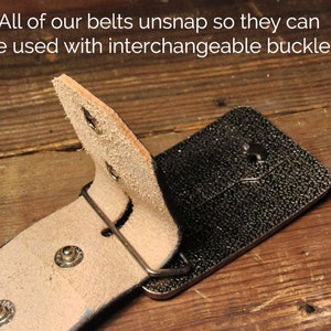 May include: A brown leather belt with a silver buckle. The belt is designed to be interchangeable with other buckles. The text "All of our belts unsnap so they can be used with interchangeable buckles" is visible in the image.