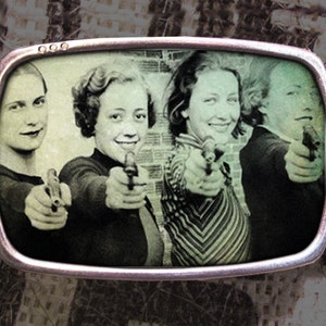 May include: A vintage black and white photograph of four people holding guns. The photo is framed in a silver belt buckle.