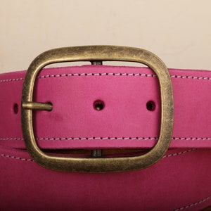 May include: A pink leather belt with a brass buckle. The belt has white stitching around the edges.