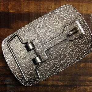 May include: A silver metal belt buckle with a textured surface. The buckle has a loop for a belt and a metal bar that slides through the loop.