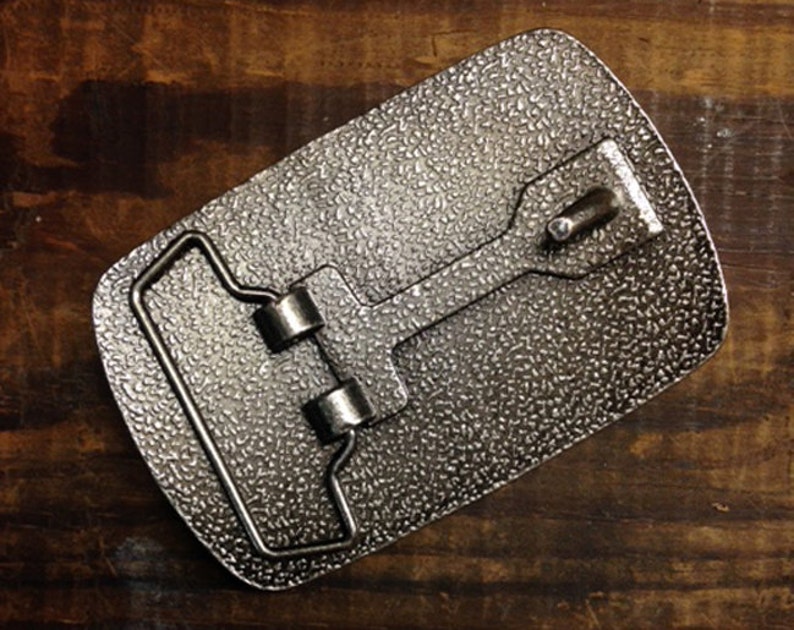 May include: A silver metal belt buckle with a textured surface. The buckle has a loop for a belt and a metal bar that slides through the loop.