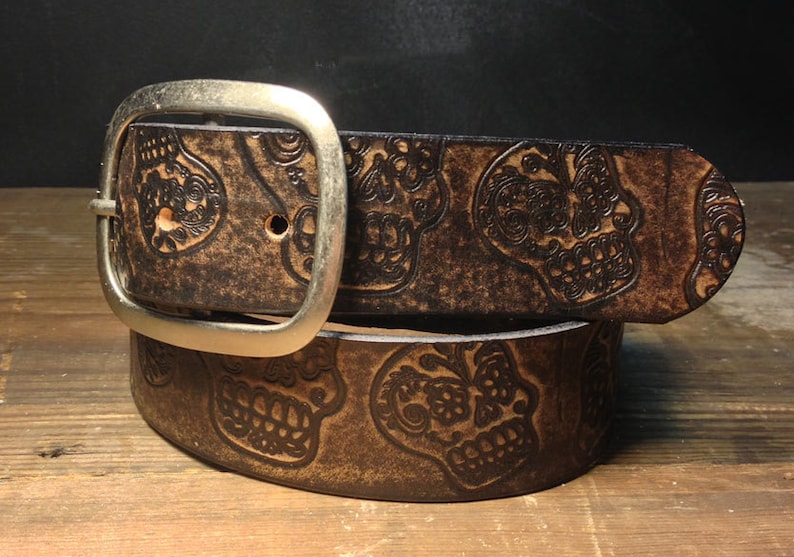May include: A brown leather belt with a silver buckle and a pattern of embossed sugar skulls.