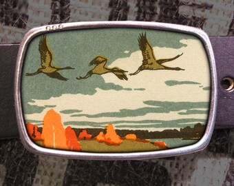 May include: A silver belt buckle featuring a vintage illustration of three birds in flight over a landscape with orange and green hills. The buckle has a textured surface and a slightly rounded shape.