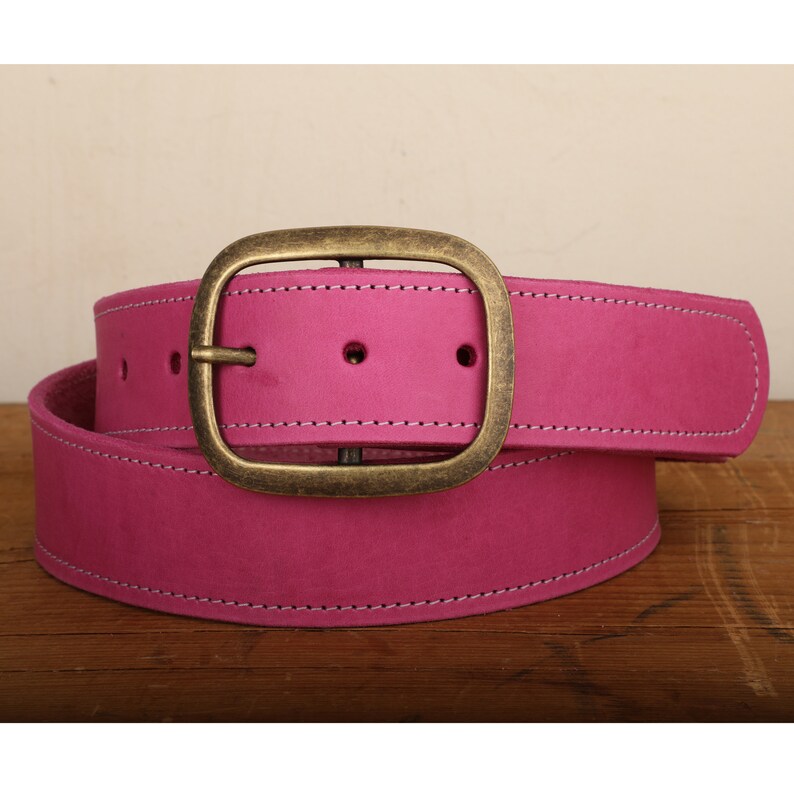 May include: A pink leather belt with a brass buckle. The belt has a stitched edge and is folded in half.