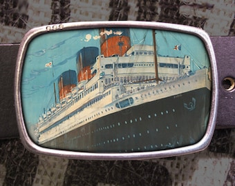 May include: A silver belt buckle featuring a vintage image of a large ocean liner. The ship is white with black accents and has a French flag flying from the mast. The ship's name, "Paris", is visible on the side.