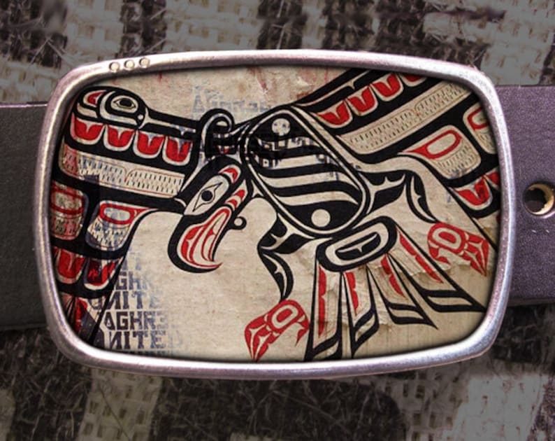 May include: A silver belt buckle with a black, red, and white design of a stylized eagle. The design is reminiscent of Native American art.