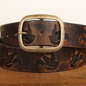 May include: A dark brown leather belt featuring an embossed pattern of flying birds throughout the strap. The belt is secured with a large, rectangular, antique brass-toned metal buckle with a rounded frame and a single prong closure mechanism.
