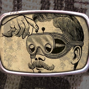 May include: A silver belt buckle with a vintage illustration of a person wearing goggles. The person has a mustache and is looking up. The buckle has a distressed, aged look.