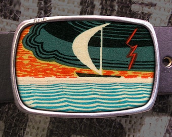 May include: A silver belt buckle featuring a colourful image of a sailboat on the water with a red lightning bolt in the sky. The image is set against a blue and green background.