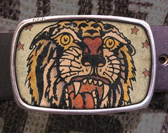 May include: A rectangular silver belt buckle featuring a detailed tiger face design with orange, yellow, and black stripes. The tiger's face is surrounded by small star shapes. The buckle is attached to a dark brown leather belt.