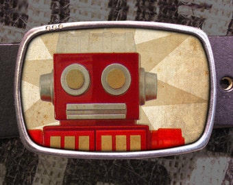 May include: A silver belt buckle with a red robot face on a vintage-style background. The robot has large round eyes and a rectangular body.