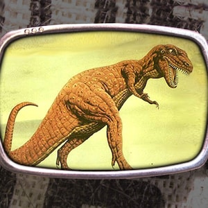 May include: A silver belt buckle with a yellow background and a brown dinosaur illustration. The dinosaur is facing right and has its mouth open.