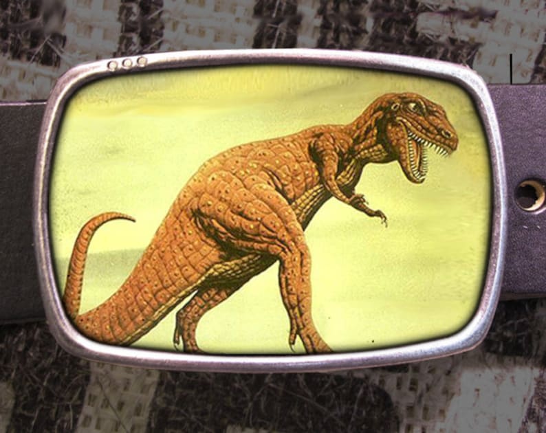 May include: A silver belt buckle with a yellow background and a brown dinosaur illustration. The dinosaur is facing right and has its mouth open.