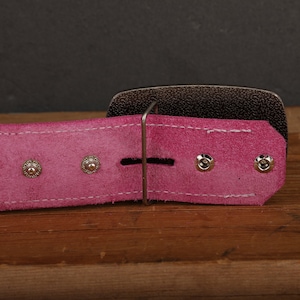 May include: Close-up of a pink leather belt with a silver buckle. The belt has three silver rivets on each side of the buckle.
