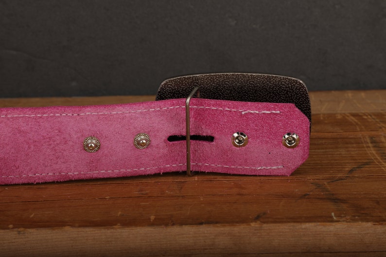 May include: Close-up of a pink leather belt with a silver buckle. The belt has three silver rivets on each side of the buckle.