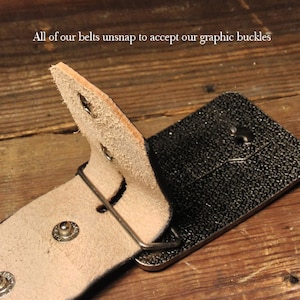 May include: A close-up of a brown leather belt with a black buckle. The belt is designed to unsnap to accept a graphic buckle. The text "All of our belts unsnap to accept our graphic buckles" is visible in the image.