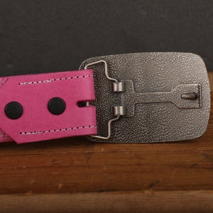 May include: A pink leather belt with a silver buckle. The buckle has a textured surface and a simple design.