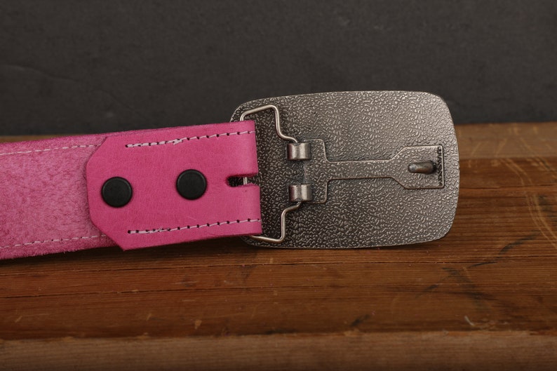 May include: A pink leather belt with a silver buckle. The buckle has a textured surface and a simple design.